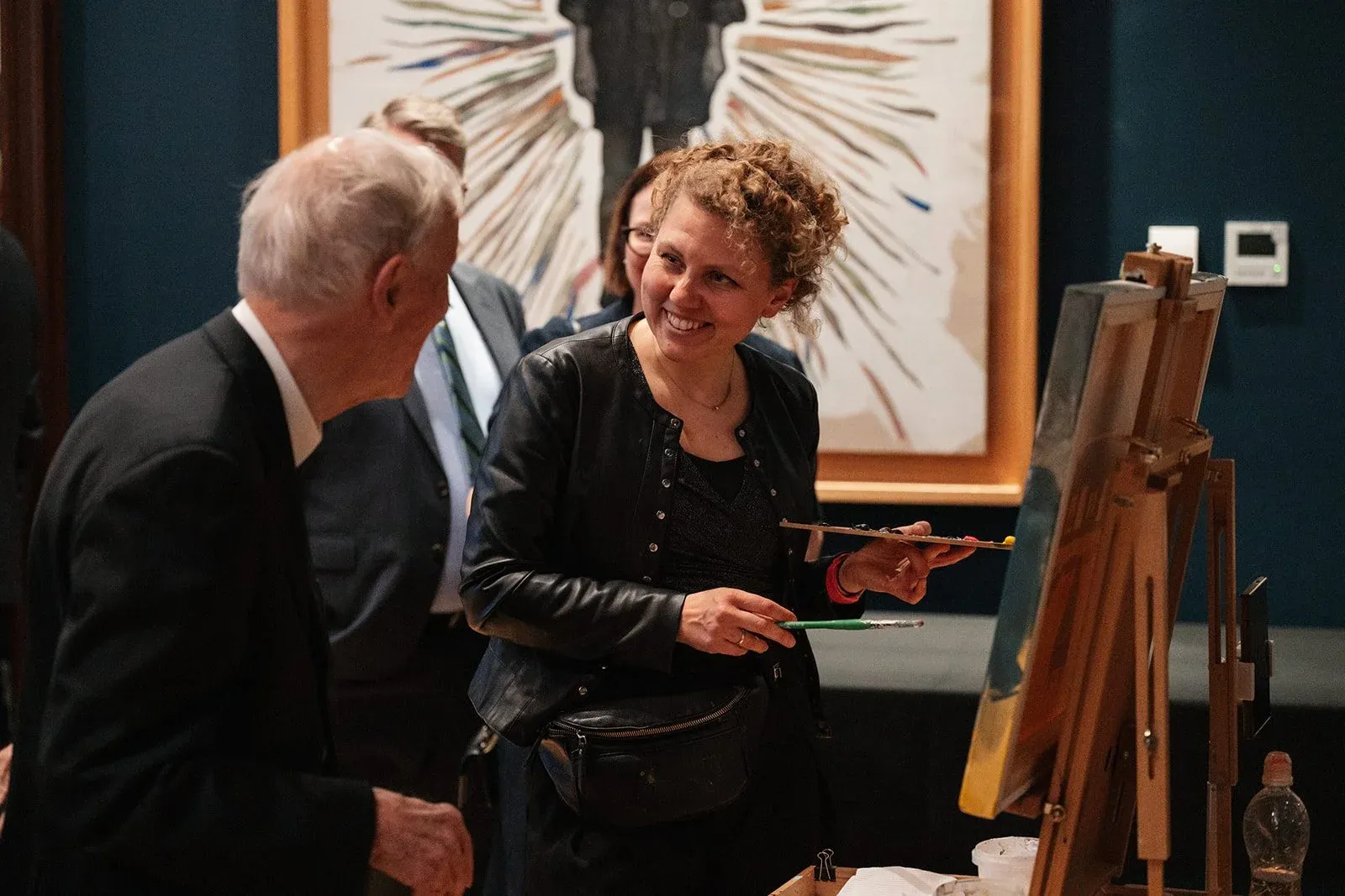 A woman smiling while holding a paint palette and brush, standing next to a man in a suit. They are at an art event with easels and paintings in the background.