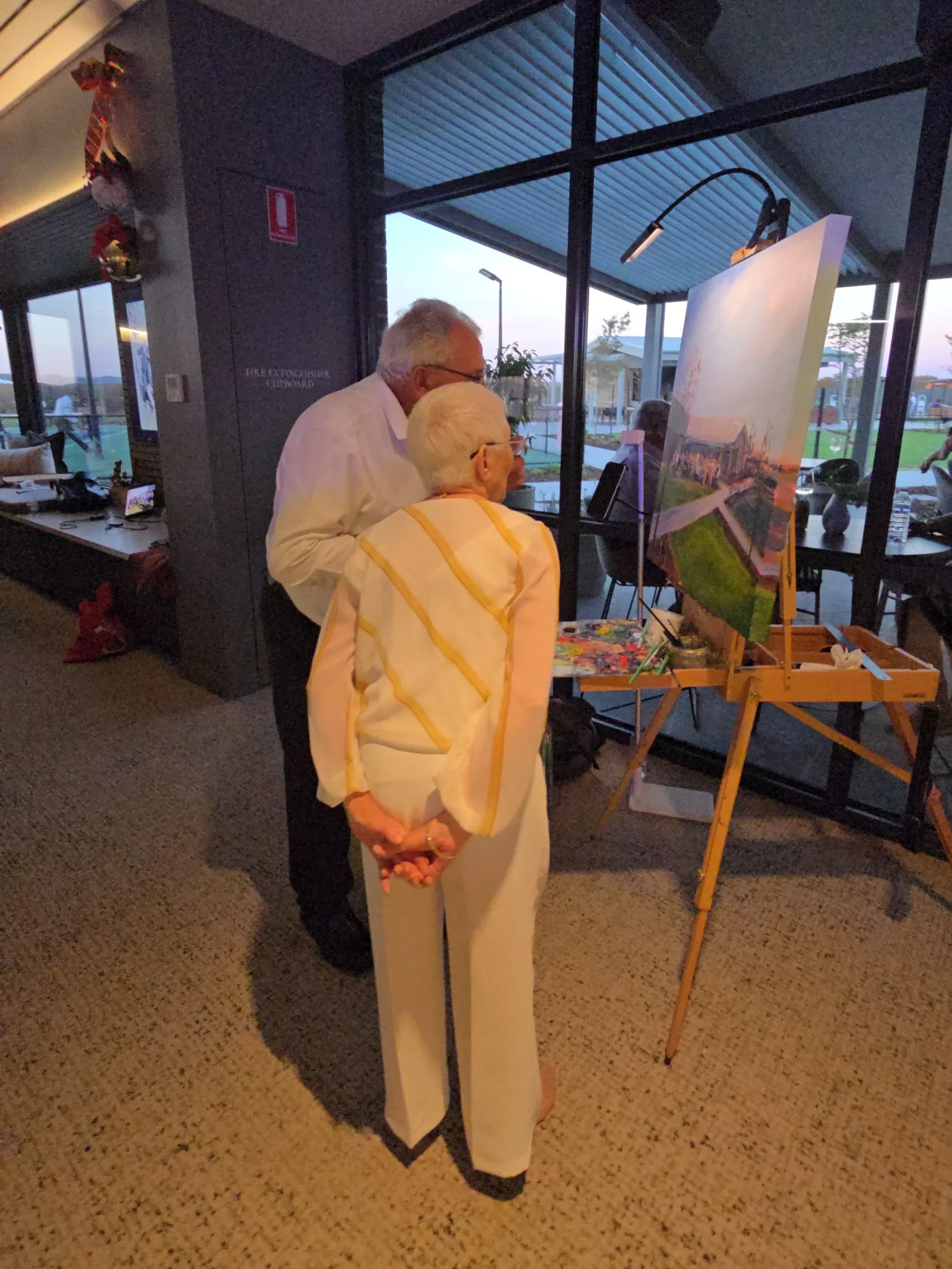 An elderly couple with white hair, one wearing a white dress with yellow stripes, are standing close together and looking at a painting on an easel inside a room with large glass windows. The room is decorated with Christmas ornaments.