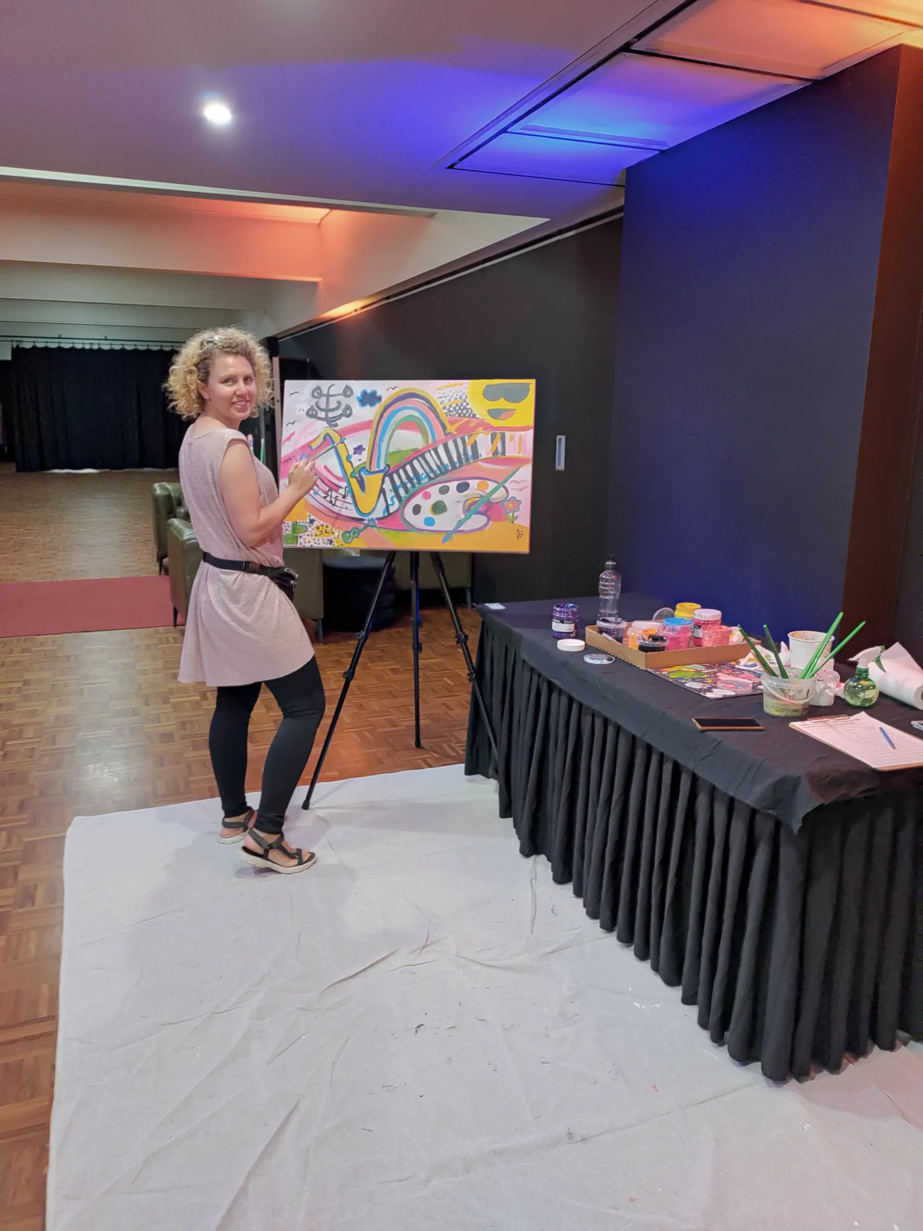 A woman with curly blonde hair wearing a pink dress and black pants, standing on a white sheet, painting a colorful rainbow on a canvas in an indoor space with dark walls and a wooden floor.
