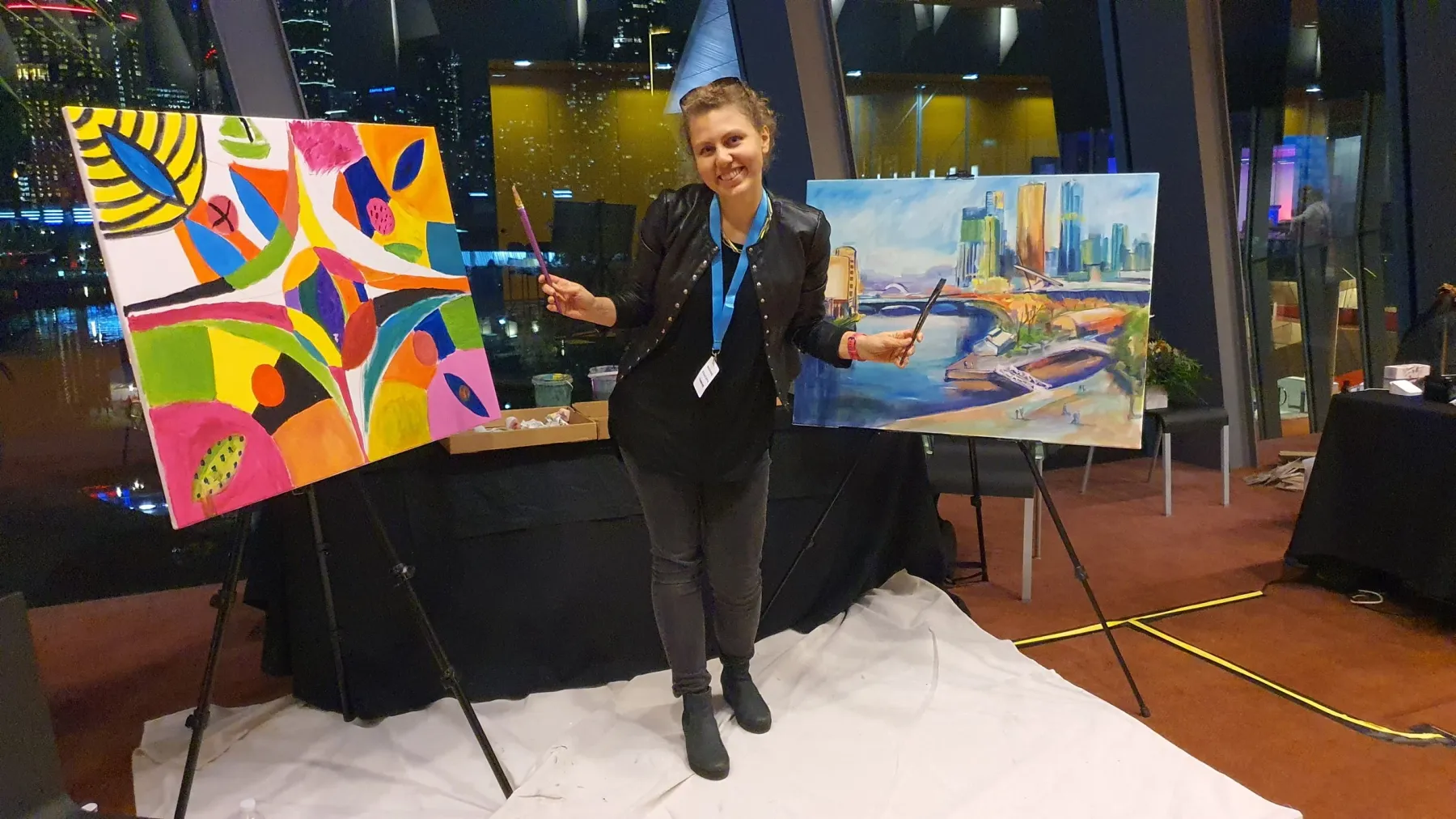 A woman smiling and holding paint brushes standing between two finished paintings at an art event, with city lights visible through the windows behind her.