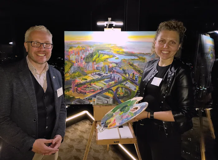A man with glasses and a woman in black leather jacket standing indoors with a colorful painting of a cityscape and river behind them. The woman holds a painter's palette and stands next to an easel.