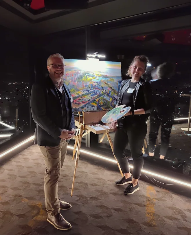 Two people standing in front of a colorful landscape painting on an easel, with a cityscape visible through large windows behind them. The man is smiling and wearing glasses, a dark jacket, and khaki pants. The woman is holding a palette and brush, w