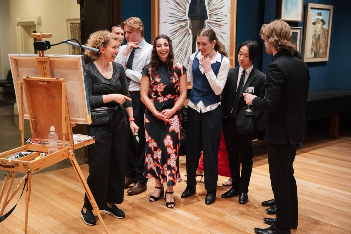 A group of people at Trinity College event. A woman with curly hair is laughing and talking to others. Several paintings hang on the dark blue wall behind them. Some people are holding drinks, and there is an easel in the foreground.