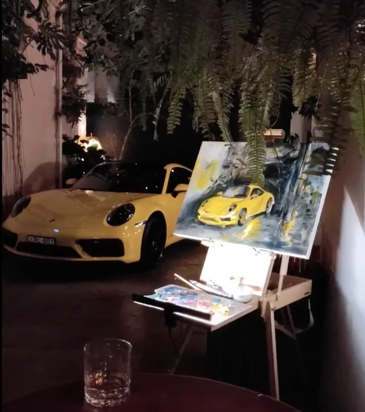 A yellow Porsche sports car parked near an easel with a painting of the same car, set outdoors at nighttime under foliage.
