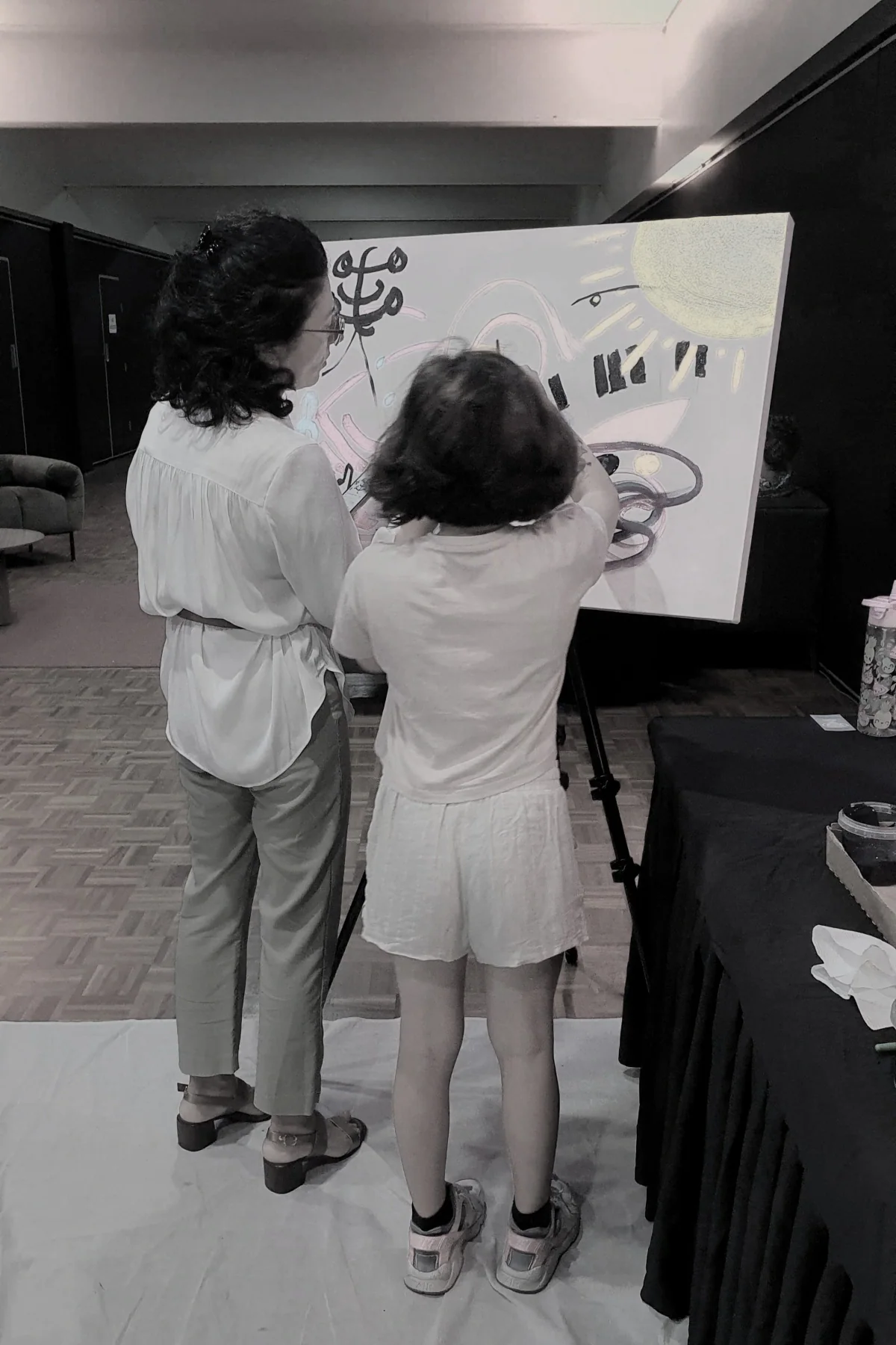 Two women standing in front of a large painting on it. They are indoors, One woman has curly hair and glasses, and the other has short hair. The floor is wood-patterned, and there is a black table with a black cloth on it to the right.