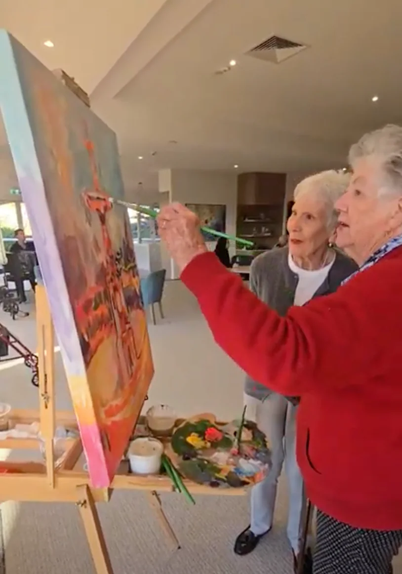 Two elderly women adding to a collaborative live painting of a colorful landscape on a canvas in a cozy indoor setting.
