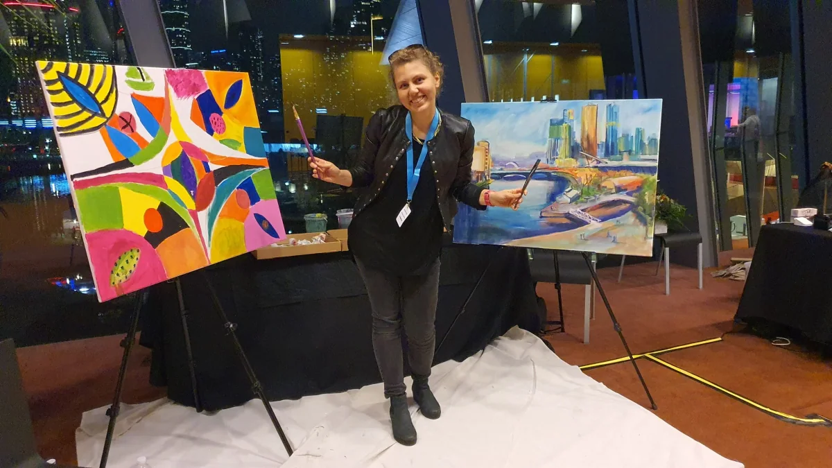 A woman smiling and holding paint brushes standing between two finished paintings at an art event, with city lights visible through the windows behind her.